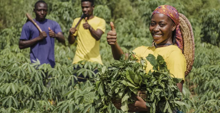 An African woman and two men are labouring in a green field, smiling at the camera with thumbs up.